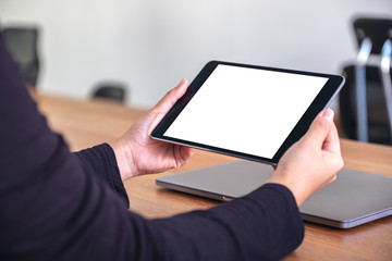 Mockup image of hands holding and using black tablet pc with blank white desktop screen with notebook on wooden table in office