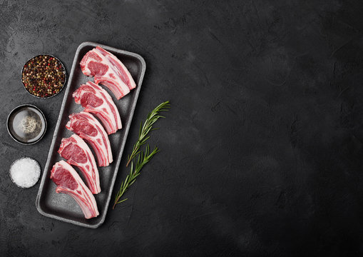 Fresh raw butchers lamb beef cutlets in plastic tray with fresh rosemary on black background.Salt, pepper and oil in steel bowl.Space for text