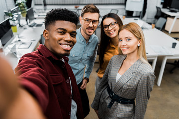 camera point of view of smiling multicultural colleagues taking selfie together in office