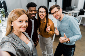 camera point of view of smiling multicultural colleagues taking selfie together in office