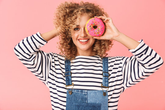 Image of content curly woman 20s wearing casual clothes eating donut while standing, isolated over pink background