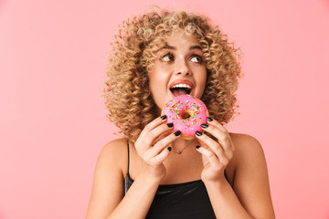 Photo of joyful curly woman 20s wearing dress eating donut while standing, isolated over pink...