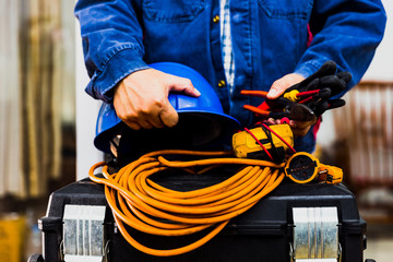 Electrian engineer holding multimeter and tools in hand, standing behind the heavy duty tool box, image including power cord, Blue hard hat (helmet) and gloves.