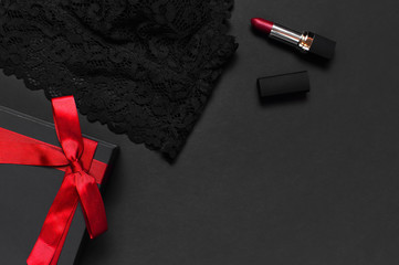 Black gift box with red ribbon, lace bra underwear, red lipstick, holographic confetti on dark background top view flat lay. Female essential erotic accessories, fashionable underwear, gift to woman