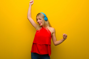 Young girl with red dress over yellow wall listening to music with headphones and dancing