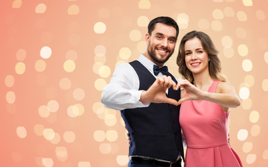 valentines day, love and people concept - happy couple in party clothes making hand heart gesture over living coral background and festive lights