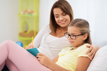 people, family and education concept - happy daughter with mother writing to notebook at home