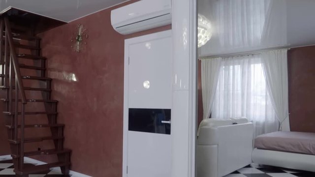 Interior of a modern living room. A white leather sofa, a white ottoman, a white wardrobe with mirrors, a white bed, a white chandelier, a white ceiling, a white air conditioner, a white curtains 