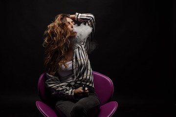 Redhead woman vaping electronic cigarette with smoke on black background closeup. Young woman smoking e-cigarette to quit tobacco. Vapor and alternative nicotine free smoking concept, copy space 