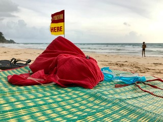 Tropical beach in Phuket island. Sand, mat for sunbathing, shoes, clothes and swimming goggles on it. The man in the background goes swimming to the sea water. Yellow-red flag rescuers "swim here".