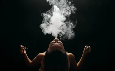 Fotobehang Redhead woman vaping electronic cigarette with smoke on black background closeup. Young woman smoking e-cigarette to quit tobacco. Vapor and alternative nicotine free smoking concept, copy space  © oes