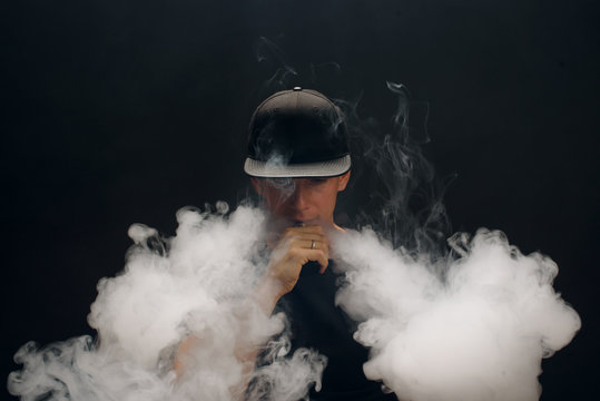 Vape man. Portrait of a handsome young white guy in a modern black cap vaping and letting off puffs of steam from an electronic cigarette