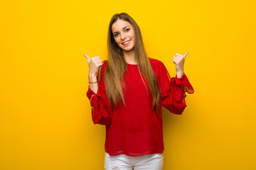 Young girl with red dress over yellow wall giving a thumbs up gesture with both hands and smiling