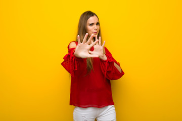 Young girl with red dress over yellow wall is a little bit nervous and scared stretching hands to the front