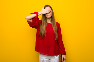 Young girl with red dress over yellow wall covering eyes by hands. Do not want to see something