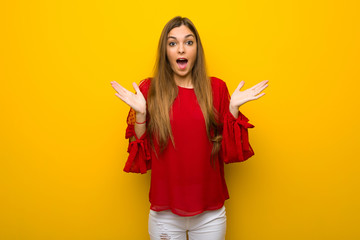 Young girl with red dress over yellow wall with surprise and shocked facial expression