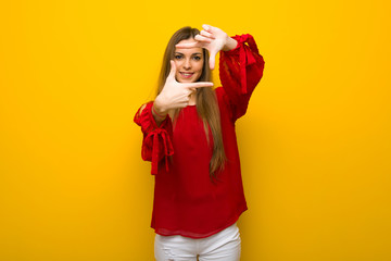 Fototapeta premium Young girl with red dress over yellow wall focusing face. Framing symbol