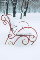 Snow-covered bench with heart symbol in the city park. Forged metal and wooden park bench and trees covered by heavy snow. Winter date, romance, Valentine's Day,