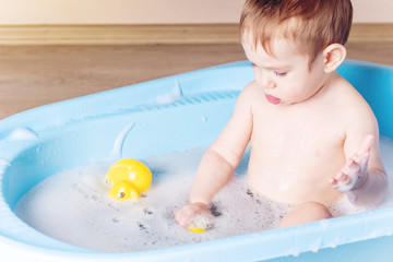 Obraz na płótnie Canvas Cute boy washing in blue bath in bathroom. Baby is playing with a yellow duck and soap foam.
