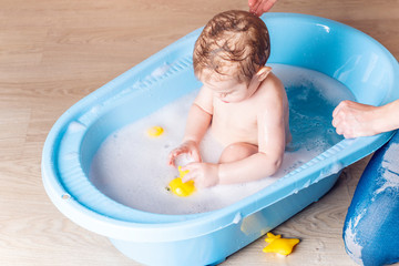 Mom washing little boy in a blue bath in the bathroom. Baby playing with a yellow duck and soap bubbles.