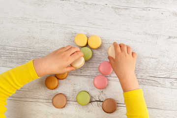 Girl's hands with yellow sleeves playing with macarons on wooden table - top view