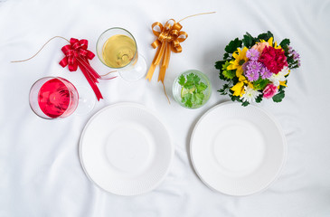Top view. Set of drinks and empty white dish on table covered by white cloth. Decorated with colorful flowers.