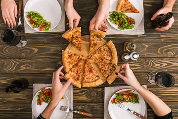 cropped view of friends sharing pizza while having dinner with salads at wooden table