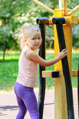 Little girl with blond hair on the sports field. The child is engaged on the street training apparatus.