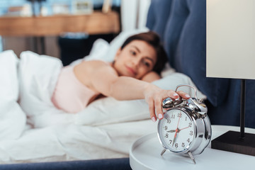 selective focus of young woman turning off alarm clock in bedroom during morning time at home