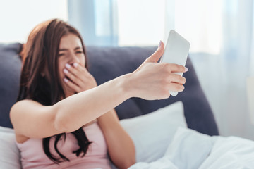 girl yawning and taking selfie on smartphone while sitting on bed during morning time at home