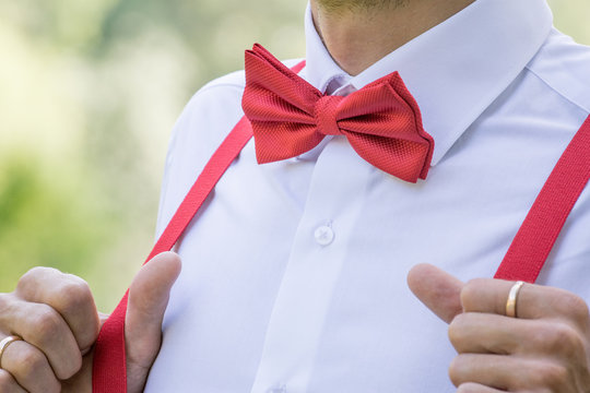 The groom in a shirt with a red bow tie and in suspenders