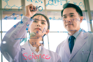 A handsome asian professor proud to teaching biology and chemist for young beautiful girl scientist to writing table of the elements on glass board in laboratory at morning classroom.