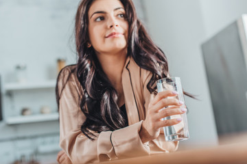 low angle view of beautiful young woman holding glass with water and sitting at wooden tabletop in kitchen at home