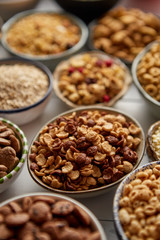 Close up and selective focus. Composition of different kinds cereals placed in ceramic bowls with cornflakes, granola, cereals and oatmeal. Flat lay, top view on white wooden table.