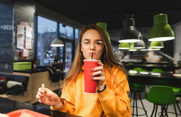 Portrait of a young girl with a red glass when sitting in the fast food restaurant is drinking a...
