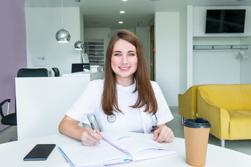 Portrait of young smiling female doctor making notes in notebook while sitting behind the table with phone and coffee inside of medical clinic. Student on practice. Healthcare and medicine. Copy space