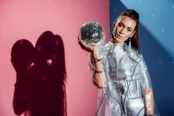 fashionable model in silver bodysuit and raincoat posing with disco ball on pink and blue background