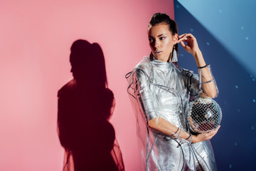 stylish young woman in metallic bodysuit and raincoat posing with disco ball on pink and blue background