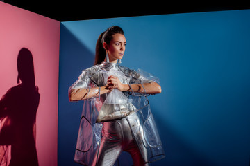 trendy young woman in silver bodysuit and raincoat holding plastic bag with fish for fashion shoot on pink and blue background