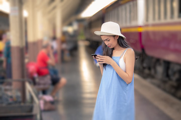 Obraz na płótnie Canvas A beautiful charming asian girl with white panama hat and light blue dress standing beside train station platform. Using smart mobile phone. Feeling look happy and relax. Portrait lady travel concept.