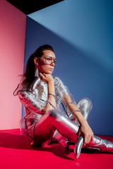 fashionable elegant woman in metallic bodysuit posing with silver bananas on pink and blue background