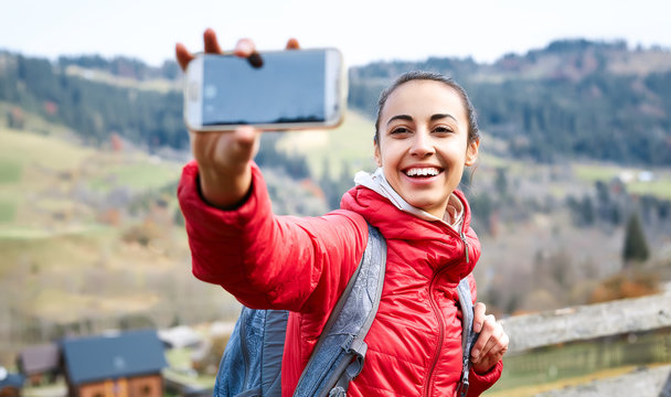 smiling happy woman hiker with backpack, wearing in red jacket and orange pants, making selfie on the woods background