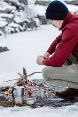 Winter camp. Man warms hands near a fire and cook coffee  the near of the frozen lake. Concept adventure active vacations outdoor. 