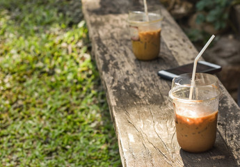 Iced coffee in plastic container placed on rustic wooden bar with green grass garden in background.