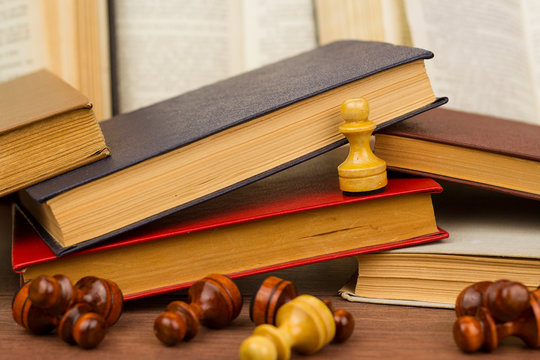 White pawn placed on stack of old book above defeated chess pieces on wooden table for concept of victory 