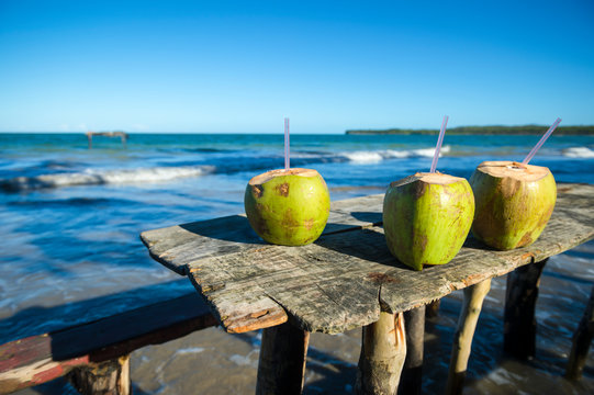 Bright scenic view of a trio of green coconuts with plastic straws resting on a rustic wooden picnic table on the beach in Bahia, Brazil