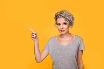 Happy surprised blonde young female smiling broadly at camera, pointing fingers away, showing something interesting and exciting on yellow studio background with copy space for your text