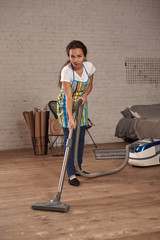 Young woman using vacuum cleaner in home living room floor, doing cleaning duties and chores, meticulous interior.