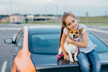 Small attractive female child embraces her favourite dog, sit together at trunk of car, have rest after stroll, enjoy summer day, have friendly relationship. Children, pets and lifestyle concept.