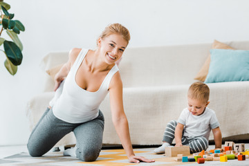 attractive woman doing stretching exercise on carpet and toddler boy playing with multicolored wooden cubes in living room